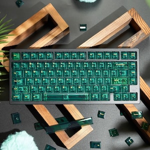 Könnte beinhalten: Eine schwarze mechanische Tastatur mit transparenten grünen Tastenkappen. Die Tastatur hat ein rechteckiges Design mit Standard-QWERTZ-Layout. Mehrere lose Tastenkappen sind um die Tastatur verstreut. Der Hintergrund umfasst dekorative Holzelemente.