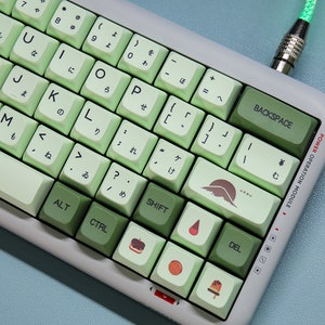May include: A green and white keyboard with Japanese characters on the keys. The keyboard has a variety of keycaps with different designs, including a mountain, a strawberry, a donut, an orange, and a popsicle. The keyboard is connected to a green cable.