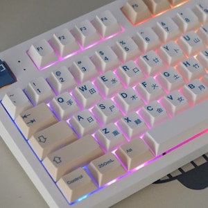 Könnte beinhalten: Eine weiße mechanische Tastatur mit cremefarbenen Tastenkappen und blauer Beschriftung. Die Tastatur hat eine regenbogenfarbene Hintergrundbeleuchtung und eine blaue Tastenkappe mit dem chinesischen Schriftzeichen für "Milch" darauf.
