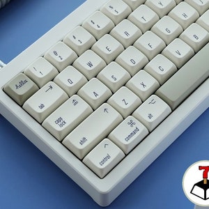 Könnte beinhalten: Eine weiße mechanische Tastatur mit cremefarbenen Tastenkappen. Die Tastatur hat eine graue Leertaste und ein Spiralkabel. Die Tastenkappen sind mit Buchstaben, Zahlen und Symbolen beschriftet. Das Wort "hello" ist auf einer Tastenkappe aufgedruckt.