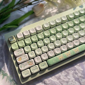 May include: A mint green mechanical keyboard with white and light green keycaps. The keyboard features custom keycaps with cartoon illustrations and decorative icons. The keyboard is designed for gaming or typing.