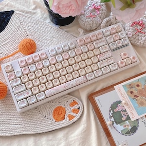 May include: A white mechanical keyboard with pastel pink and cream keycaps featuring a floral design and cat paw prints. The keyboard has a white and pink floral themed keycap set and a white and pink floral themed wrist rest.