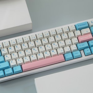 Könnte beinhalten: Eine weiße mechanische Tastatur mit einem rosa-blauen Farbschema. Die Tasten sind mit japanischen Schriftzeichen und englischen Buchstaben bedruckt. Die Tastatur hat ein rosa-blaues Farbschema.