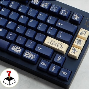 May include: A navy blue mechanical keyboard with white keycaps featuring space-themed illustrations, including stars, planets, comets, and a telescope. The keyboard has a number row, function keys, and arrow keys.