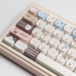 Könnte beinhalten: Eine rosa Tastatur mit weißen Tasten und benutzerdefinierten Tastenkappen mit niedlichen Katzenillustrationen. Die Tastenkappen sind in einem Pastellfarbschema mit braunen, rosa und blauen Akzenten gehalten. Die Tastatur hat eine Leertaste mit einem weißen und rosa Design.