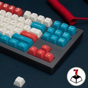 May include: A black and gray mechanical keyboard with red and blue keycaps. The keyboard has a number pad and arrow keys. The keyboard is on a blue surface. A white keycap with a red dot is in the foreground.
