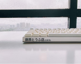 May include: A white mechanical keyboard with off-white keycaps. The keyboard has Japanese text on the front. The background shows a window with a cityscape and rain.