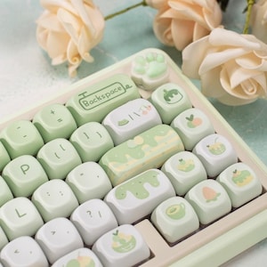Könnte beinhalten: Eine grün-weiße Tastatur mit benutzerdefinierten Tastenkappen, die verschiedene Designs zum Thema Essen zeigen, darunter einen grünen Smoothie, einen Pfirsich und eine Kiwi. Die Tastatur hat einen hellgrünen Rahmen und weiße Tasten mit schwarzer Beschriftung. Die Tastenkappen sind aus glattem, glänzendem Kunststoff.