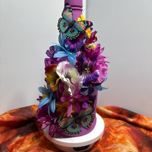 Pu&ograve; includere: Una bottiglia decorativa viola adornata con fiori artificiali colorati e due farfalle. La bottiglia &egrave; decorata con fiori viola, blu, gialli e bianchi. Le farfalle hanno ali turchesi e rosa.