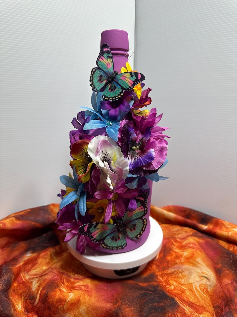 Pu&ograve; includere: Una bottiglia decorativa viola adornata con fiori artificiali colorati e due farfalle. La bottiglia &egrave; decorata con fiori viola, blu, gialli e bianchi. Le farfalle hanno ali turchesi e rosa.