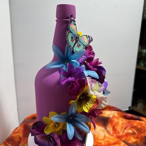 Pu&ograve; includere: Una bottiglia viola decorata con fiori artificiali colorati e una farfalla. I fiori includono fiori blu, viola, gialli e bianchi. La farfalla ha un corpo turchese e ali rosa e gialle. La bottiglia &egrave; su un supporto bianco.