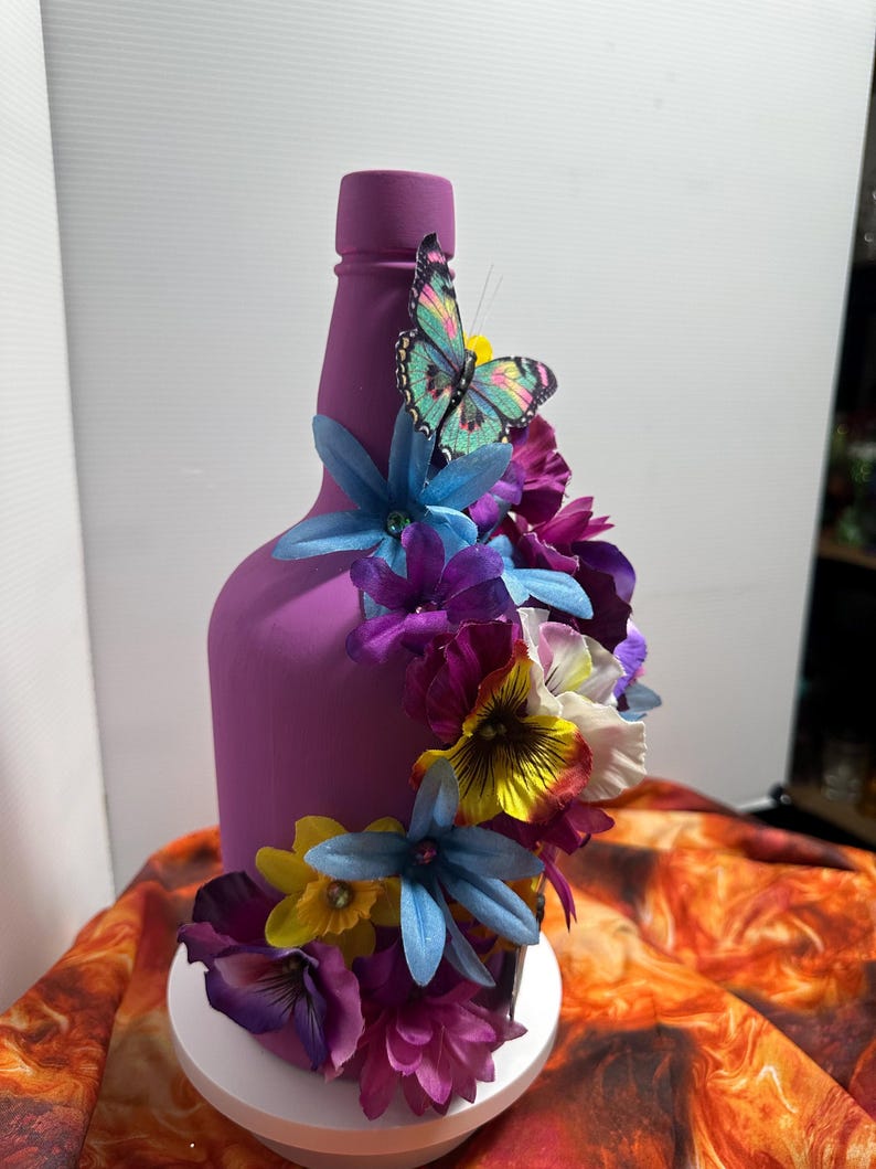 Pu&ograve; includere: Una bottiglia viola decorata con fiori artificiali colorati e una farfalla. I fiori includono fiori blu, viola, gialli e bianchi. La farfalla ha un corpo turchese e ali rosa e gialle. La bottiglia &egrave; su un supporto bianco.