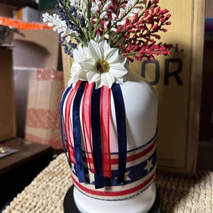 Op de afbeelding: Een witte keramische vaas versierd met rode, witte en blauwe linten en een band met sterrenpatroon. De vaas is gevuld met witte, rode en blauwe bloemstukken, wat een patriottisch thema cre&euml;ert. De vaas is ongeveer 25 cm hoog.