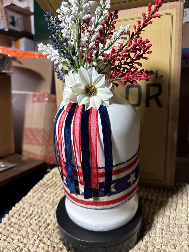 Op de afbeelding: Een witte keramische vaas versierd met rode, witte en blauwe linten en een band met sterrenpatroon. De vaas is gevuld met witte, rode en blauwe bloemstukken, wat een patriottisch thema cre&euml;ert. De vaas is ongeveer 25 cm hoog.