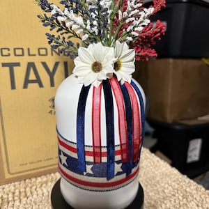 Op de afbeelding: Een witte vaas versierd met rood, wit en blauw lint en een ontwerp van een sterrenbanner. De vaas is gevuld met rood, wit en blauw bloemstukken en witte madeliefjes accenten. De vaas staat op een zwarte basis.