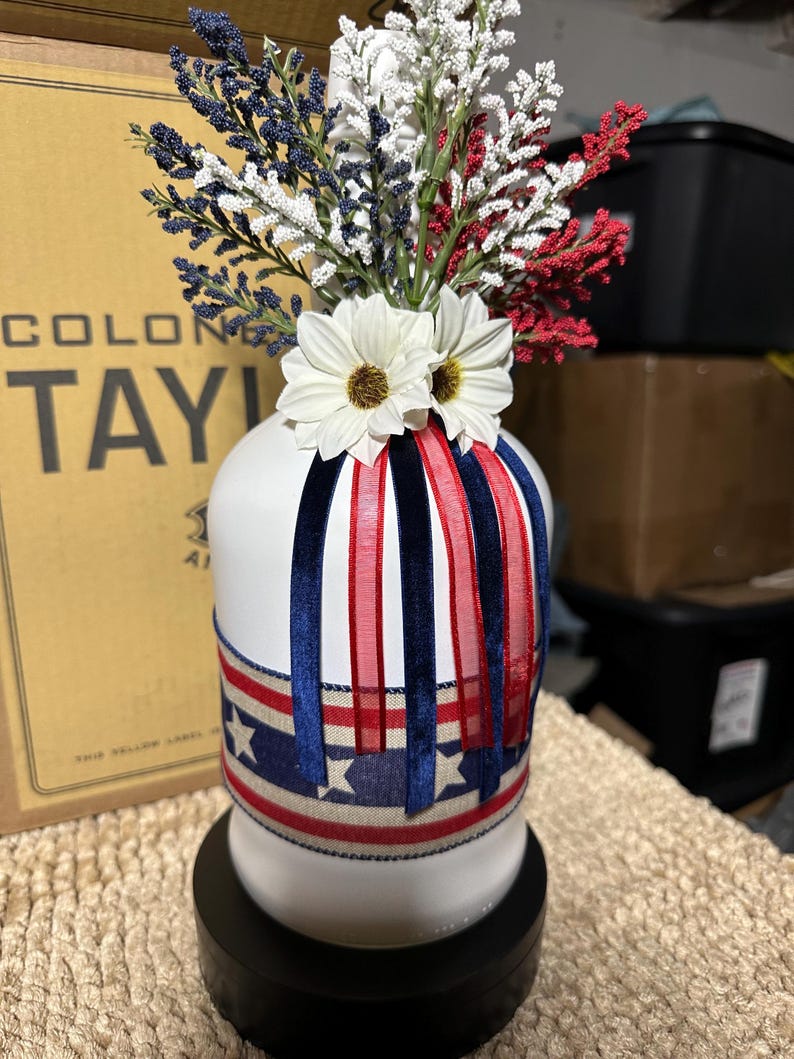 Op de afbeelding: Een witte vaas versierd met rood, wit en blauw lint en een ontwerp van een sterrenbanner. De vaas is gevuld met rood, wit en blauw bloemstukken en witte madeliefjes accenten. De vaas staat op een zwarte basis.