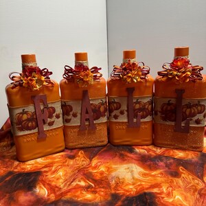 May include: Four orange bottles decorated with the word "FALL" in individual letters. Each bottle has a burlap band with pumpkin graphics, topped with orange and red floral arrangements and a cork. The bottles are on a fall-themed fabric.