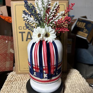 Op de afbeelding: Een witte keramische vaas versierd met rode, witte en blauwe linten en bloemaccenten. De vaas is versierd met een patriottisch thema, met een mix van kunstbloemen en decoratieve linten. De vaas staat op een zwarte voet.
