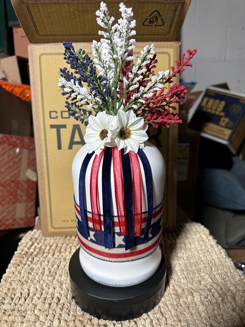 Op de afbeelding: Een witte keramische vaas versierd met rode, witte en blauwe linten en bloemaccenten. De vaas is versierd met een patriottisch thema, met een mix van kunstbloemen en decoratieve linten. De vaas staat op een zwarte voet.