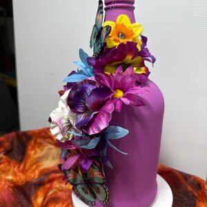 Pu&ograve; includere: Una bottiglia dipinta di viola adornata con fiori artificiali colorati e farfalle. La composizione include fiori viola, gialli e blu, con farfalle che aggiungono un tocco stravagante. La bottiglia &egrave; su un supporto bianco.