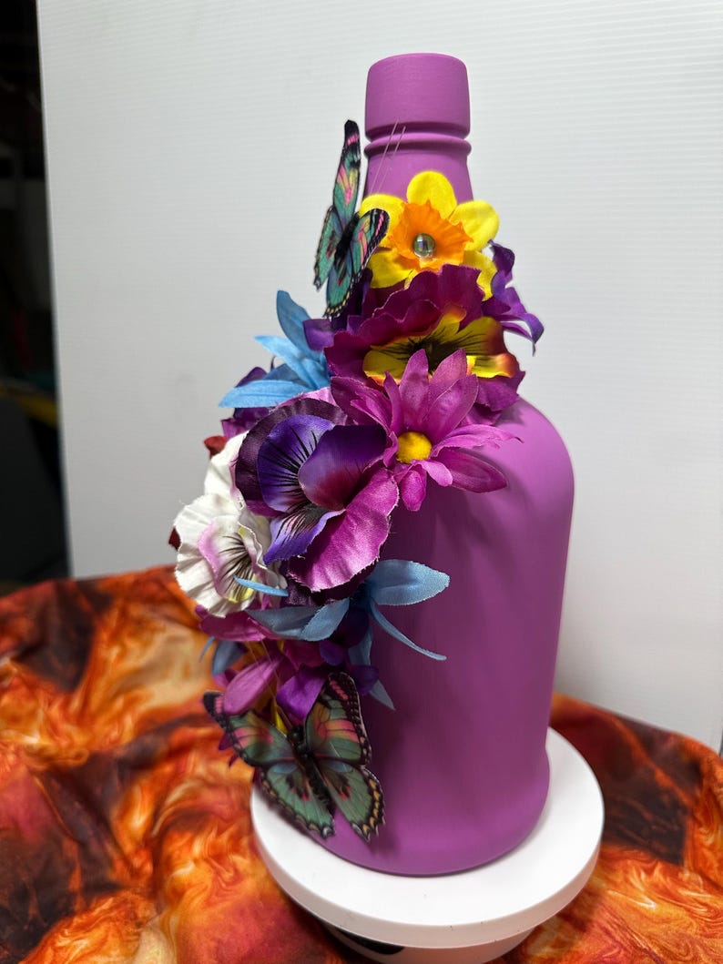 Pu&ograve; includere: Una bottiglia dipinta di viola adornata con fiori artificiali colorati e farfalle. La composizione include fiori viola, gialli e blu, con farfalle che aggiungono un tocco stravagante. La bottiglia &egrave; su un supporto bianco.