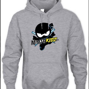 Könnte beinhalten: Grauer Kapuzenpullover mit einem schwarzen Ninja-Motiv, weißen Augen und blauen Akzenten. Der Text "NINJA KIDZ tv" ist darunter abgebildet. Der Hoodie hat einen Kordelzug und eine Kängurutasche.