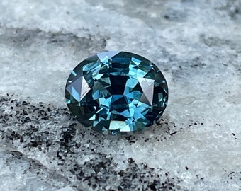 May include: A teal blue oval-shaped gemstone with a faceted cut. The gemstone is sitting on a white and gray textured surface.