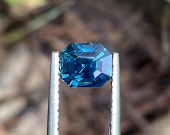 May include: A deep blue gemstone with a square emerald cut, held in a pair of tweezers. The gemstone is faceted and has a brilliant shine.