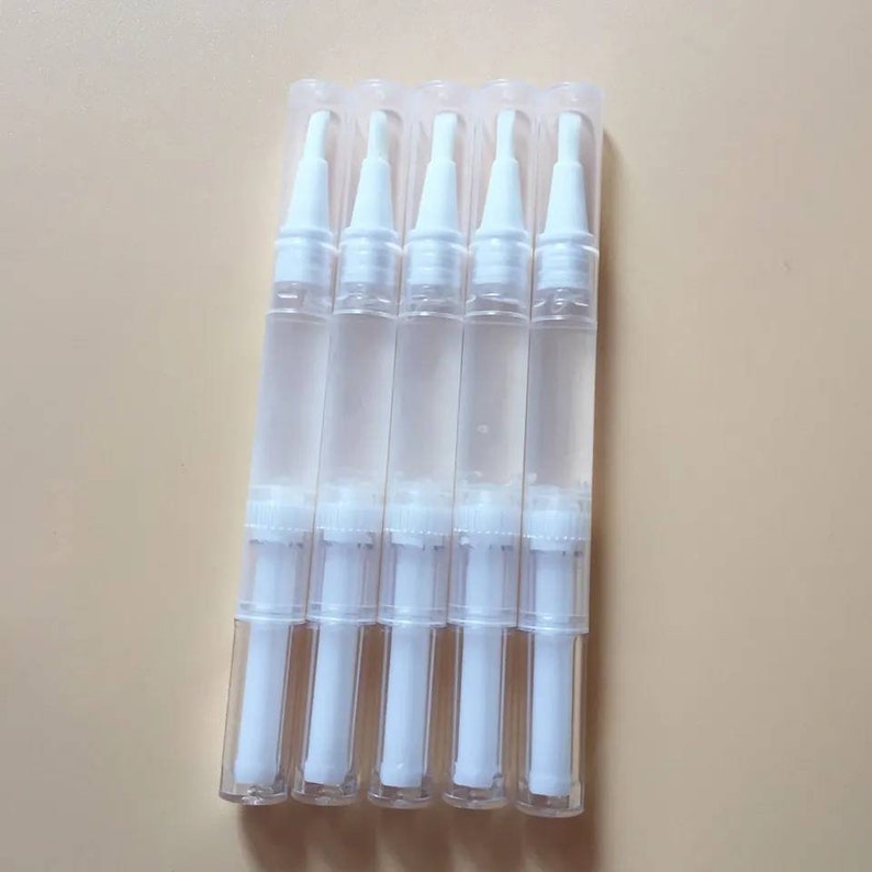 May include: Five clear nail treatment pens with white applicator tips are arranged vertically. The pens are filled with a clear liquid and are packaged in a clear plastic blister pack. The background is a solid, light beige colour.