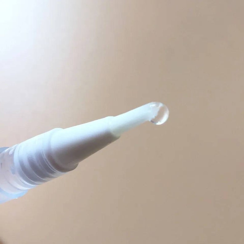 May include: A close-up shot of a white applicator with a clear liquid droplet at the tip. The applicator is cylindrical and made of plastic. The background is a plain, light beige colour.