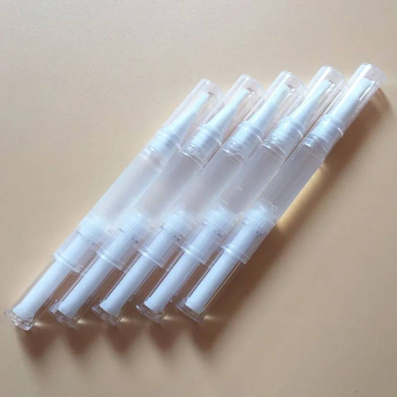 May include: Six clear, pen-shaped applicators with white brush tips and clear caps are arranged diagonally. The image shows a beauty product, possibly for nails or skincare, against a pale beige background.