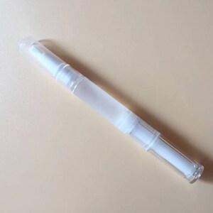 May include: A clear, pen-shaped nail care product with a white applicator tip and a frosted body. The product is designed for nail care and is shown on a light beige background.