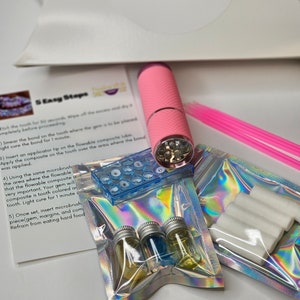 May include: A kit for applying a tooth gem, including a pink UV light, a small container of clear flowable composite, a container of small gems, a container of clear liquid, a container of blue liquid, a container of yellow liquid, a container of white cotton swabs, and a set of instructions with the text "5 Easy Steps".