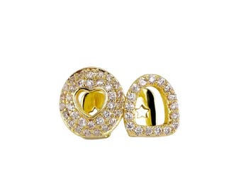 May include: A pair of gold-colored dental grills. One grill features a heart-shaped cutout, while the other has a star-shaped cutout. Both are encrusted with small, sparkling stones, creating a luxurious and eye-catching design.