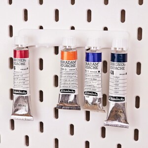 May include: Four tubes of gouache paint in a white holder. The tubes are labeled "Schmincke Designers Gouache" and "Schmincke Horadam Gouache". The colors are red, orange, blue, and indigo.