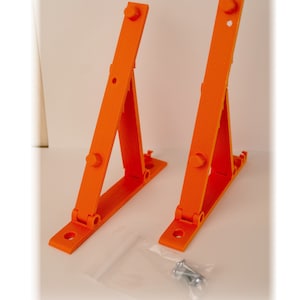 Könnte beinhalten: Zwei orangefarbene Kunststoff-Regalhalterungen mit dreieckiger Form und einem Scharnier. Die Halterungen sind für die Montage an einer Wand konzipiert und können zur Unterstützung von Regalen verwendet werden. Die Halterungen werden mit Schrauben zur Installation geliefert.