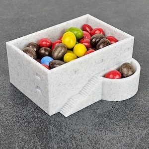 Könnte beinhalten: Ein weißer, gesprenkelter rechteckiger Behälter mit eingebauter Treppe und abgerundetem Seitenfach. Der Behälter ist mit bunten Süßigkeiten gefüllt, darunter rote, gelbe, braune, grüne und blaue.