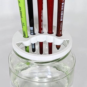 May include: A clear glass jar filled with water, featuring a white plastic brush holder. The holder contains several paint brushes in various colours and sizes, ready for use. The brushes include green, black, and red.