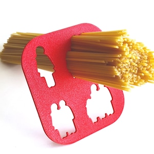 Könnte beinhalten: Ein rotes Spaghetti-Messwerkzeug mit drei ausgeschnittenen Figuren, das ein Bündel ungekochter Spaghetti hält. Das Werkzeug dient zum Abmessen von Portionen. Die Spaghetti sind hellgelb.