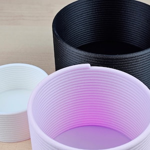 Könnte beinhalten: Drei zylindrische Behälter, die in 3D gedruckt wurden, in verschiedenen Größen. Der größte Behälter ist schwarz, der mittlere Behälter ist rosa und der kleinste Behälter ist weiß.