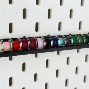 Könnte beinhalten: Ein schwarzer Fadenrollenhalter mit einer Vielzahl farbiger Fadenrollen. Die Spulen sind in den Farbtönen Weiß, Rosa, Rot, Lila, Schwarz, Blau, Türkis, Grün und Gold gehalten. Der Halter ist an einer weißen Lochwand befestigt.