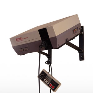 May include: A vintage Nintendo Entertainment System (NES) console in grey and beige with a black controller hanging from the power cord. The NES is mounted on a black wall mount. The console has the text "Nintendo Entertainment System" and "EUROPEAN VERSION" printed on the front.