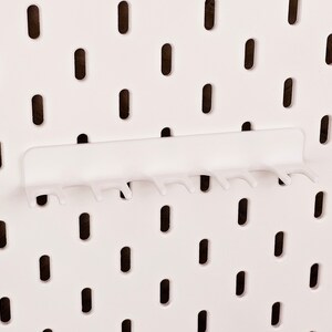 May include: White pegboard with a white plastic hook rack attached. The hook rack has seven hooks.