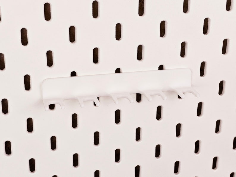 May include: White pegboard with a white plastic hook rack attached. The hook rack has seven hooks.