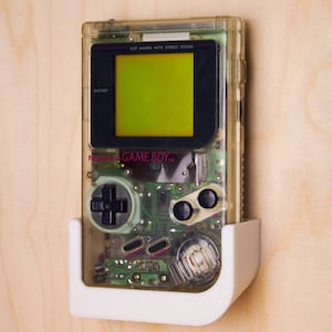 Könnte beinhalten: Eine transparente, durchsichtige Game Boy Handheld-Videospielkonsole mit einem grünen Bildschirm und dem Text "DOT MATRIX WITH STEREO SOUND" und "Nintendo GAME BOY TM" auf der Vorderseite. Die Konsole ist an einer weißen Wandhalterung befestigt.