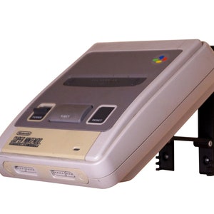 Könnte beinhalten: Eine weiße Super Nintendo Entertainment System Konsole mit einer schwarzen Wandhalterung. Die Konsole hat einen grauen Kartenschlitz und einen Ein-/Ausschalter, einen Auswurftaste und einen Reset-Knopf.