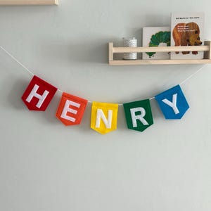 Puede incluir: Una colorida pancarta de fieltro que deletrea "HENRY" en letras blancas, ensartada en una cuerda blanca. Cada letra est&aacute; en una bandera de diferente color: rojo, naranja, amarillo, verde y azul. La pancarta est&aacute; colgada en una pared gris claro.