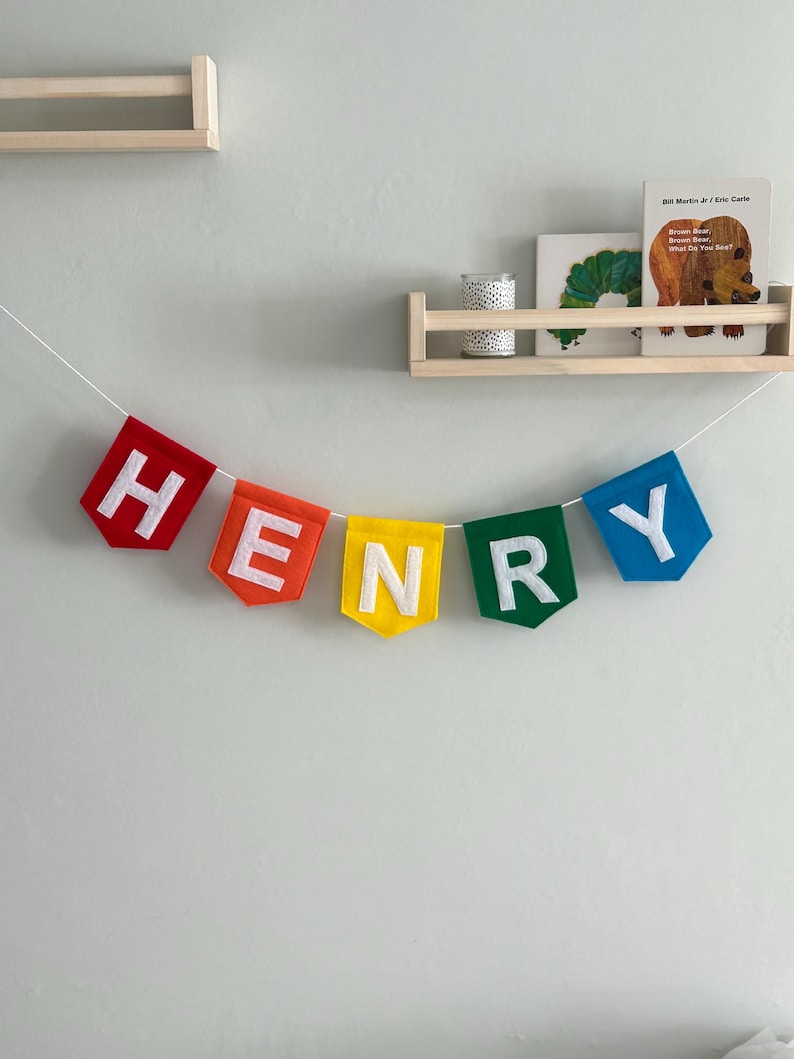 Puede incluir: Una colorida pancarta de fieltro que deletrea "HENRY" en letras blancas, ensartada en una cuerda blanca. Cada letra est&aacute; en una bandera de diferente color: rojo, naranja, amarillo, verde y azul. La pancarta est&aacute; colgada en una pared gris claro.