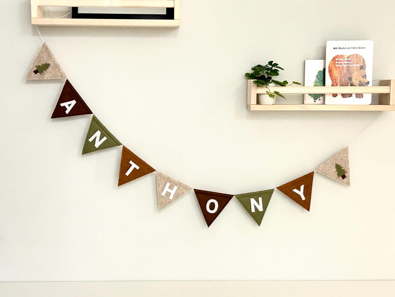 Puede incluir: Guirnalda de fieltro con el nombre "ANTHONY" en banderines triangulares. Los banderines son de color marr&oacute;n, verde y beige, con letras blancas. Dos banderines tienen un peque&ntilde;o dise&ntilde;o de &aacute;rbol. La guirnalda cuelga de una pared blanca.