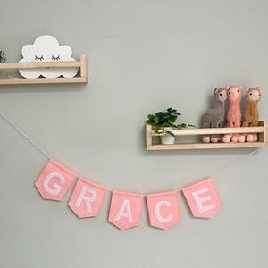 Puede incluir: Una pancarta de fieltro rosa con la palabra "GRACE" cuelga debajo de dos estantes de madera. El estante superior contiene una decoraci&oacute;n blanca en forma de nube con los ojos cerrados. El estante inferior muestra llamas de peluche.